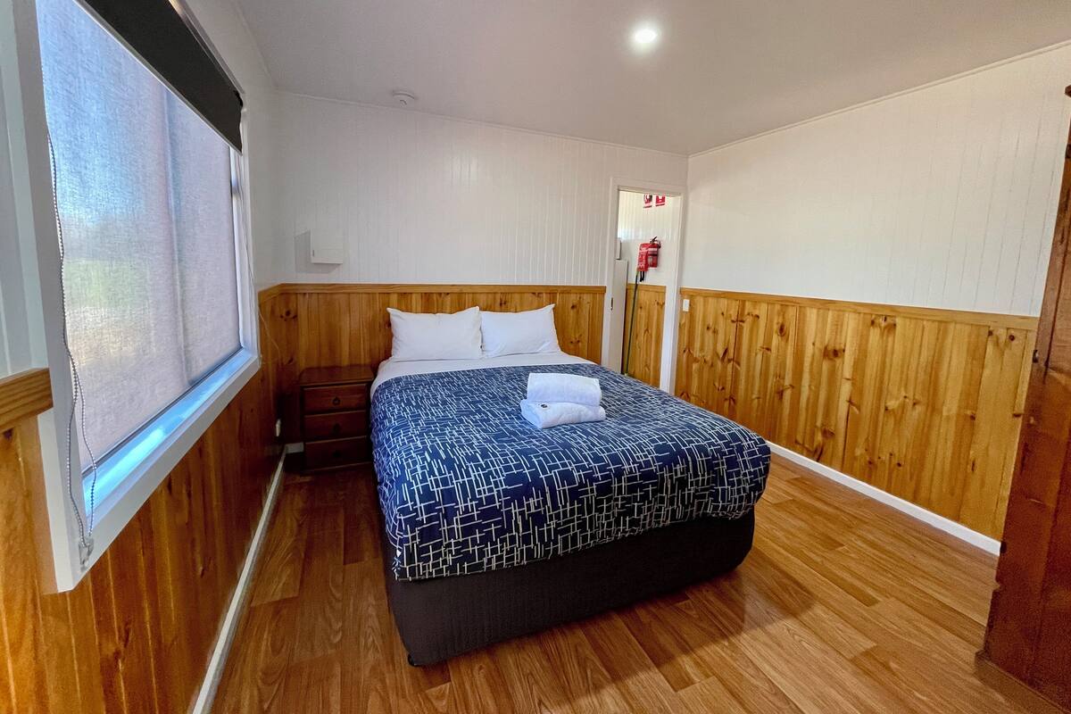 Une chambre avec un lit doté d’une tête de lit en bois, une fenêtre avec des stores, une table de chevet et un extincteur fixé au mur.