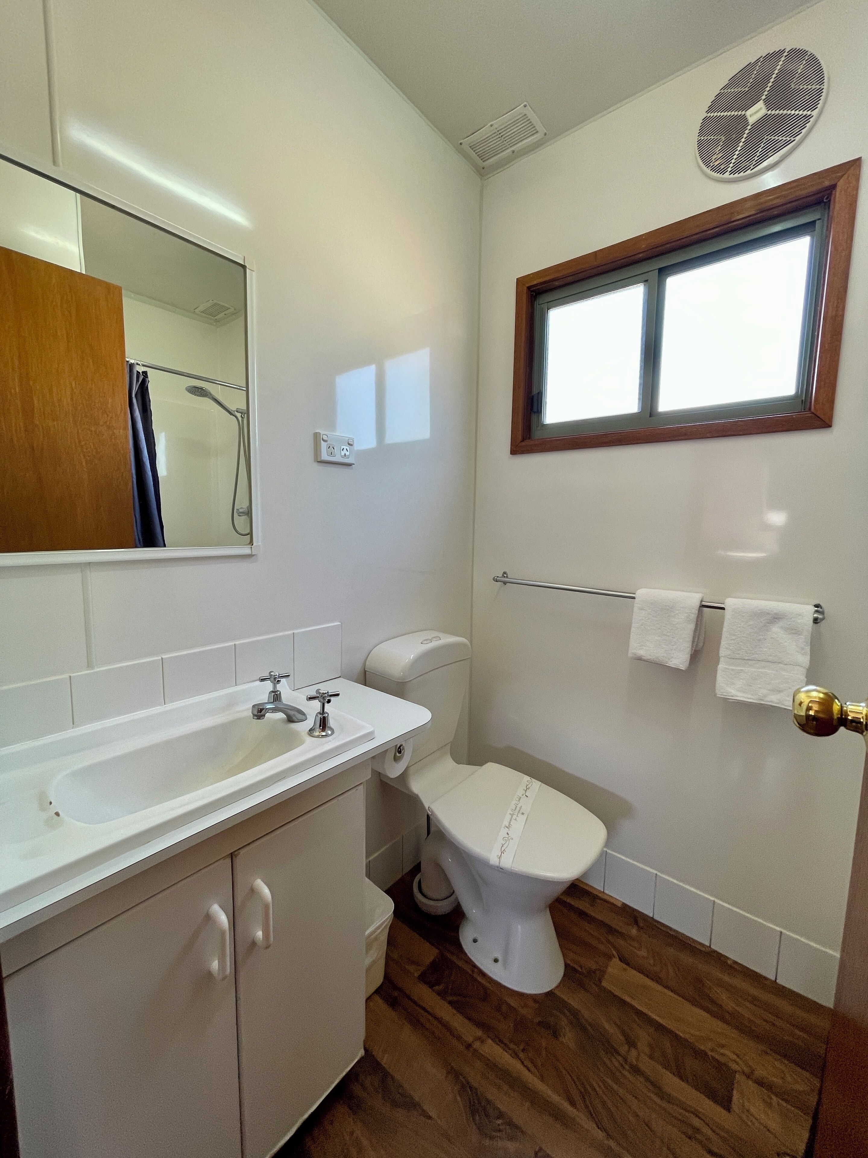 Une salle de bain avec un lavabo, des toilettes et une douche, tous de couleur blanche. Il y a une fenêtre au-dessus des toilettes et un porte-serviettes avec des serviettes.