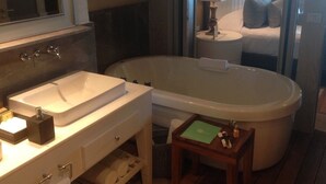Ein Badezimmer mit einer Badewanne, einem Waschbecken und einem Spiegel. Es gibt einen kleinen Tisch mit einer Lampe und einem Buch.