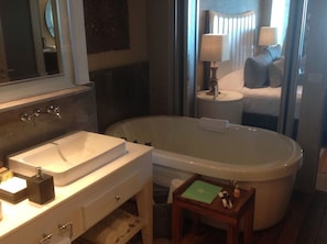 Ein Badezimmer mit einer Badewanne, einem Waschbecken und einem Spiegel. Es gibt einen kleinen Tisch mit einer Lampe und einem Buch.