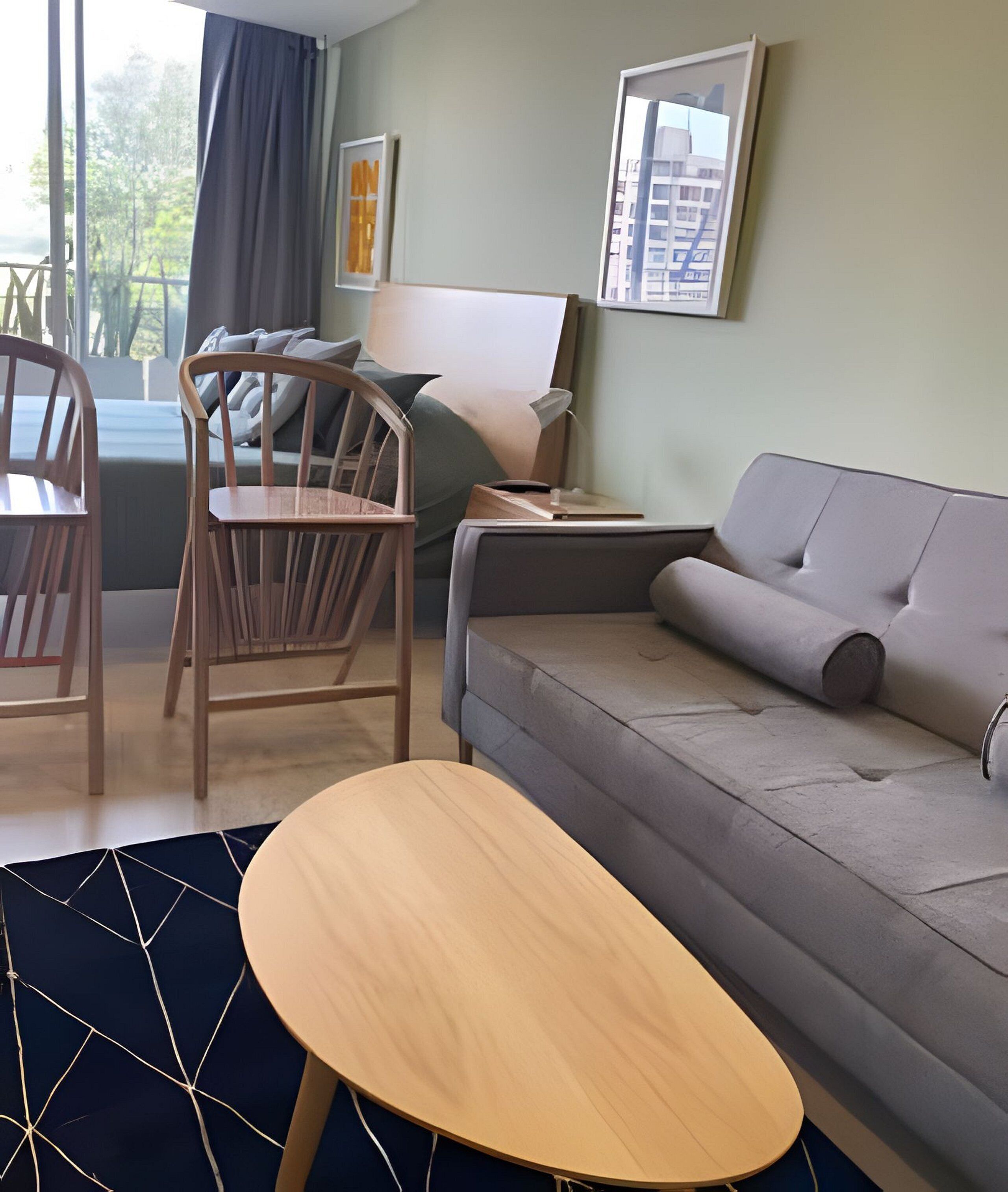 Un salon moderne avec un canapé gris, des chaises en bois, une table basse ronde et une vue sur les immeubles par la fenêtre.