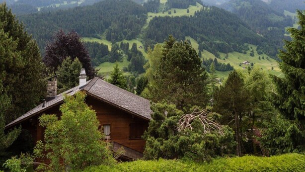 Una baita di legno con tetto spiovente, immersa in una fitta foresta con montagne sullo sfondo.