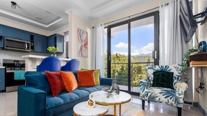 A modern living room with a blue sofa, a round coffee table, and a chair with a leafy pattern. A kitchen with dark blue cabinets and a microwave is visible in the background. - The Blooming Blaze W/ Pool (Kingston)