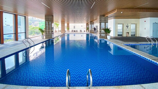 An indoor swimming pool with a wooden ceiling, clear glass walls, and a view of the surrounding area. - Peace Hotel Ha Long (Bai Chay)