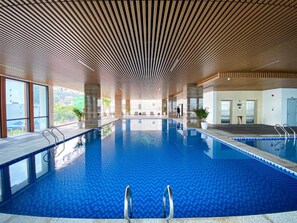 An indoor swimming pool with a wooden ceiling, clear glass walls, and a view of the surrounding area. - Peace Hotel Ha Long (Bai Chay)