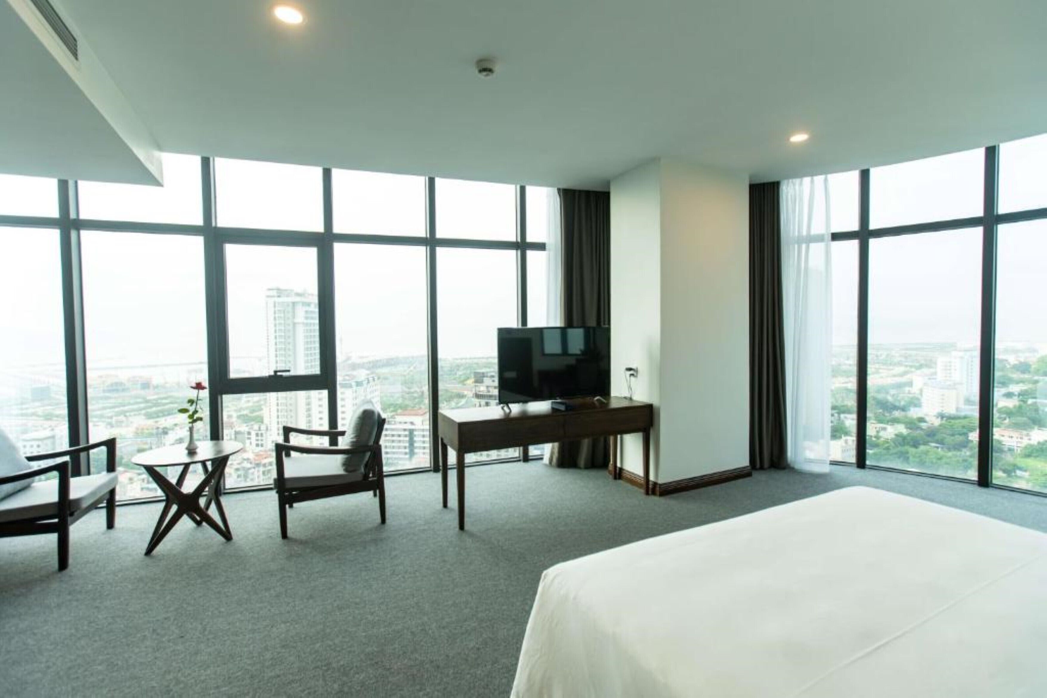 A hotel room with a large bed, a desk, a chair, a small table with a vase, and a TV.