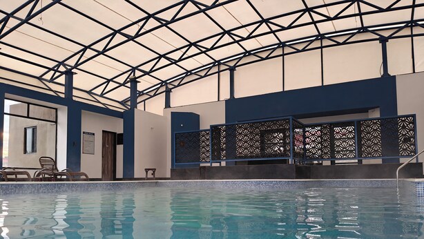 An indoor swimming pool with clear water, a metal roof structure, and a poolside area with chairs and a small table. - Tathastu Satpura (Shahpur)