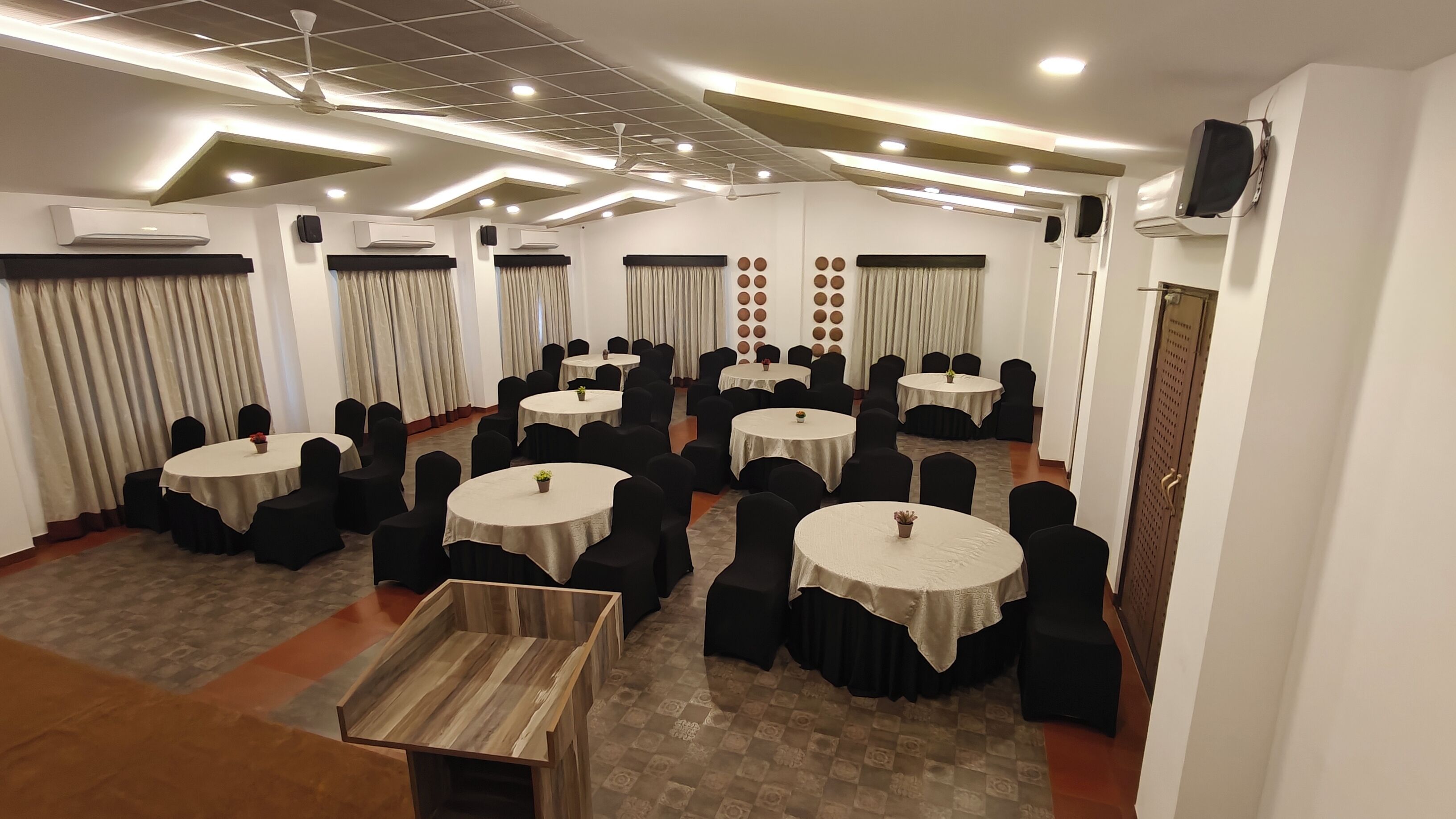A conference room with multiple round tables and black chairs, air conditioning units on the walls, and a wooden bench in the center.