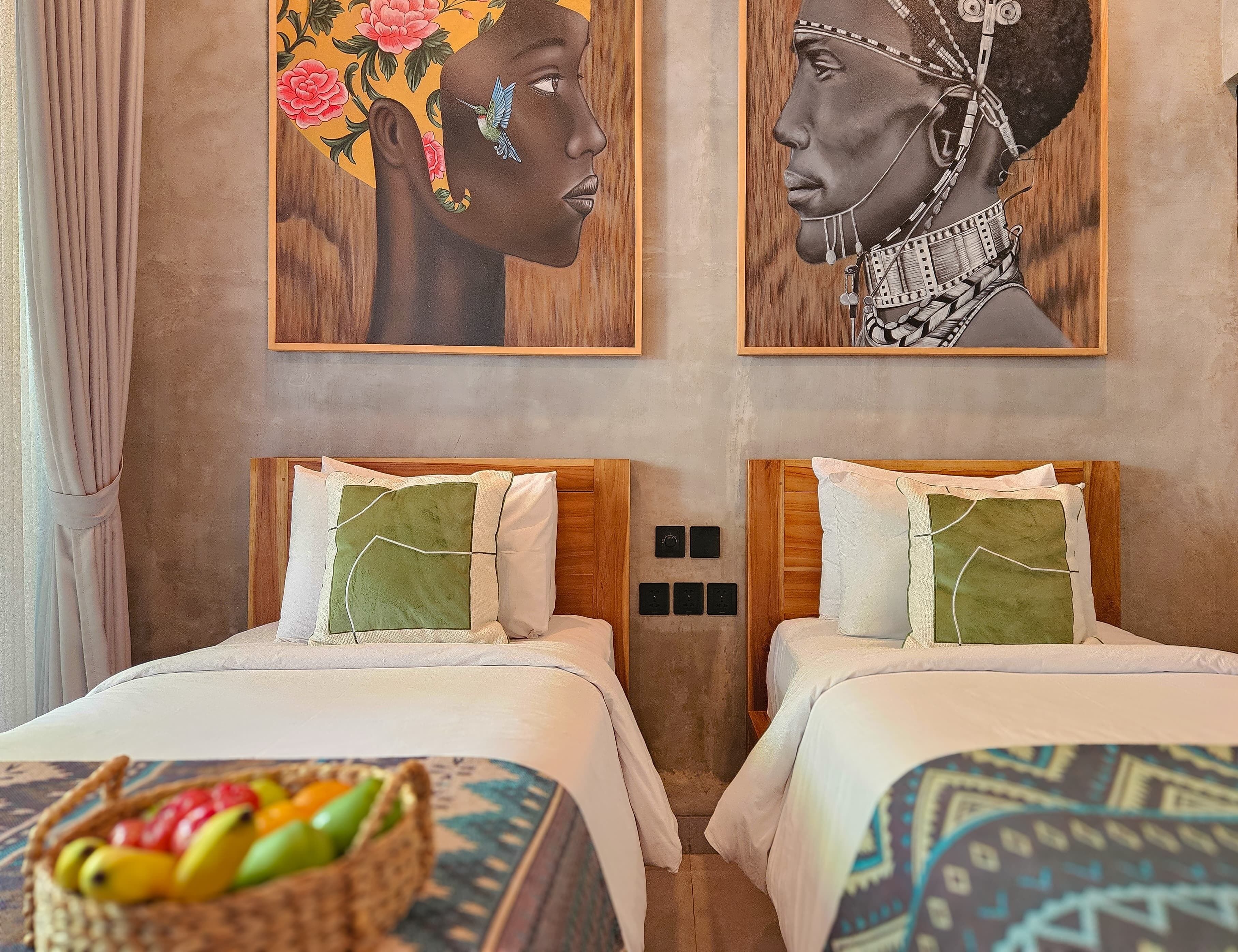 Une chambre d’hôtel avec deux lits, chacun doté d’une tête de lit en bois et de linge de lit blanc. On trouve un panier de fruits sur le sol et deux tableaux accrochés au mur au-dessus des lits.