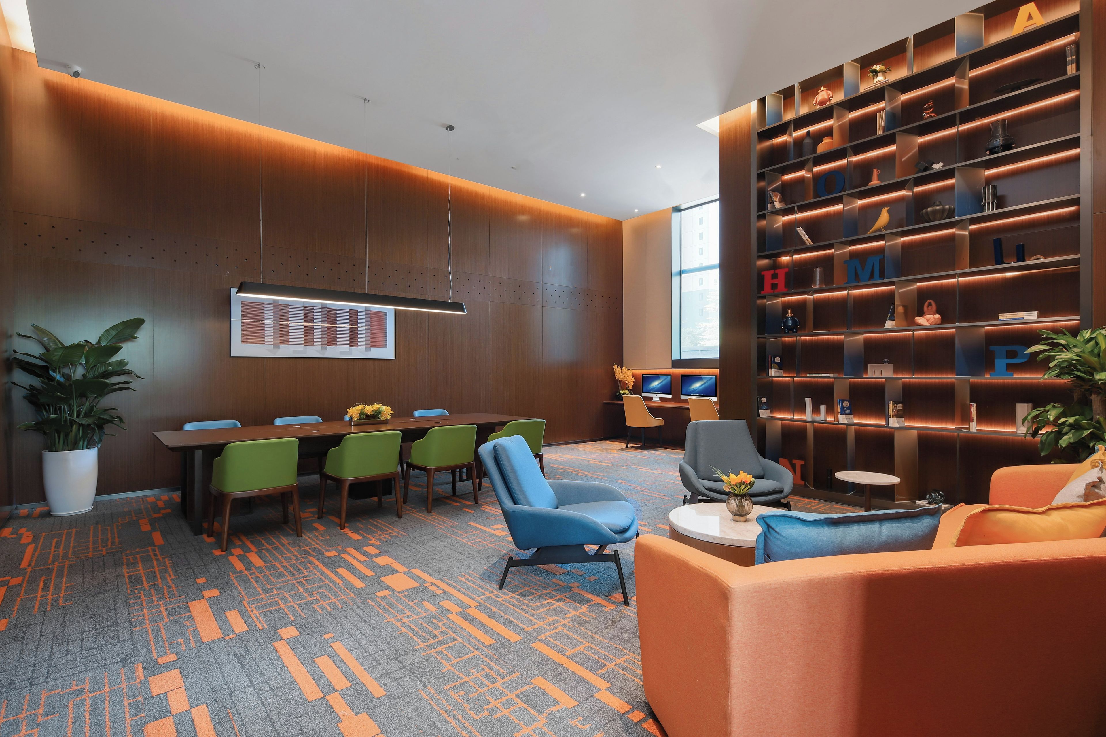 Une salle de conférence moderne, dotée d’une longue table, de chaises vertes et bleues, d’un canapé et d’une bibliothèque agrémentée d’objets décoratifs.