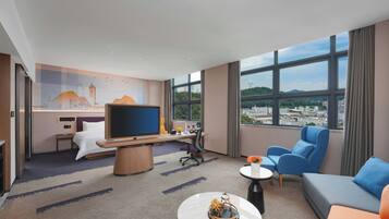 A hotel room with a bed, a desk with a computer, a TV, a sofa, and a view of a cityscape.