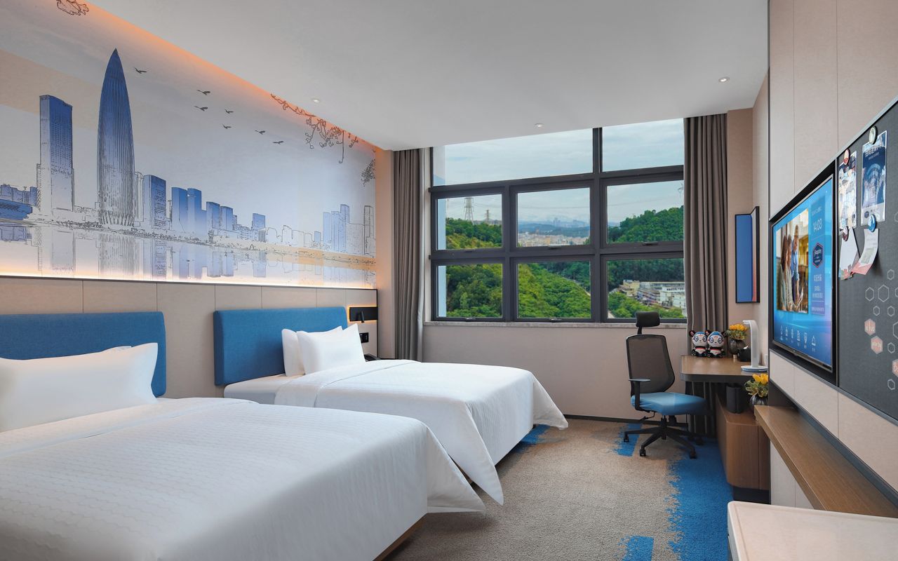 A hotel room with two beds, a desk with a computer, a TV, and a large window with a cityscape view.