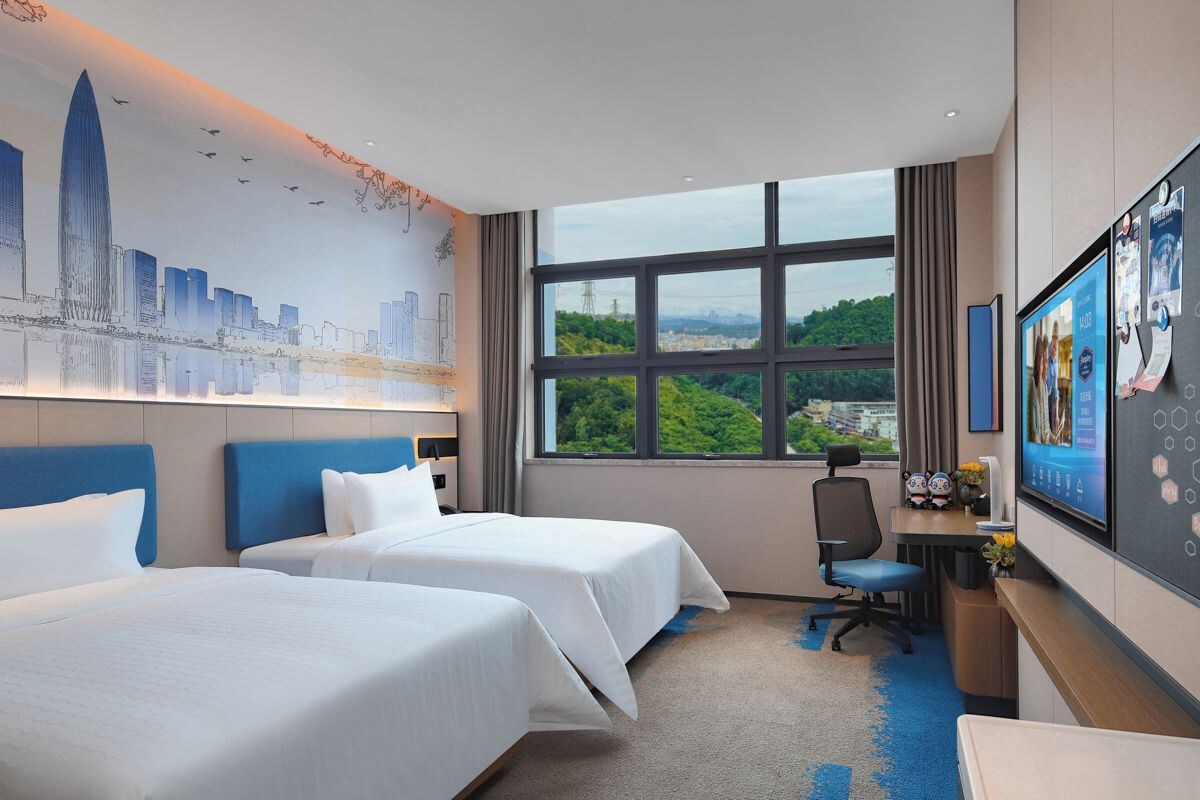 A hotel room with two beds, a desk with a computer, a TV, and a large window with a cityscape view.