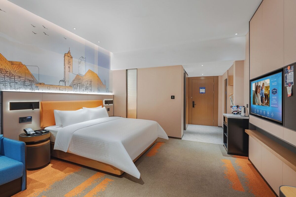 A modern hotel room with a large bed, a desk, a television, and a cityscape mural on the wall.