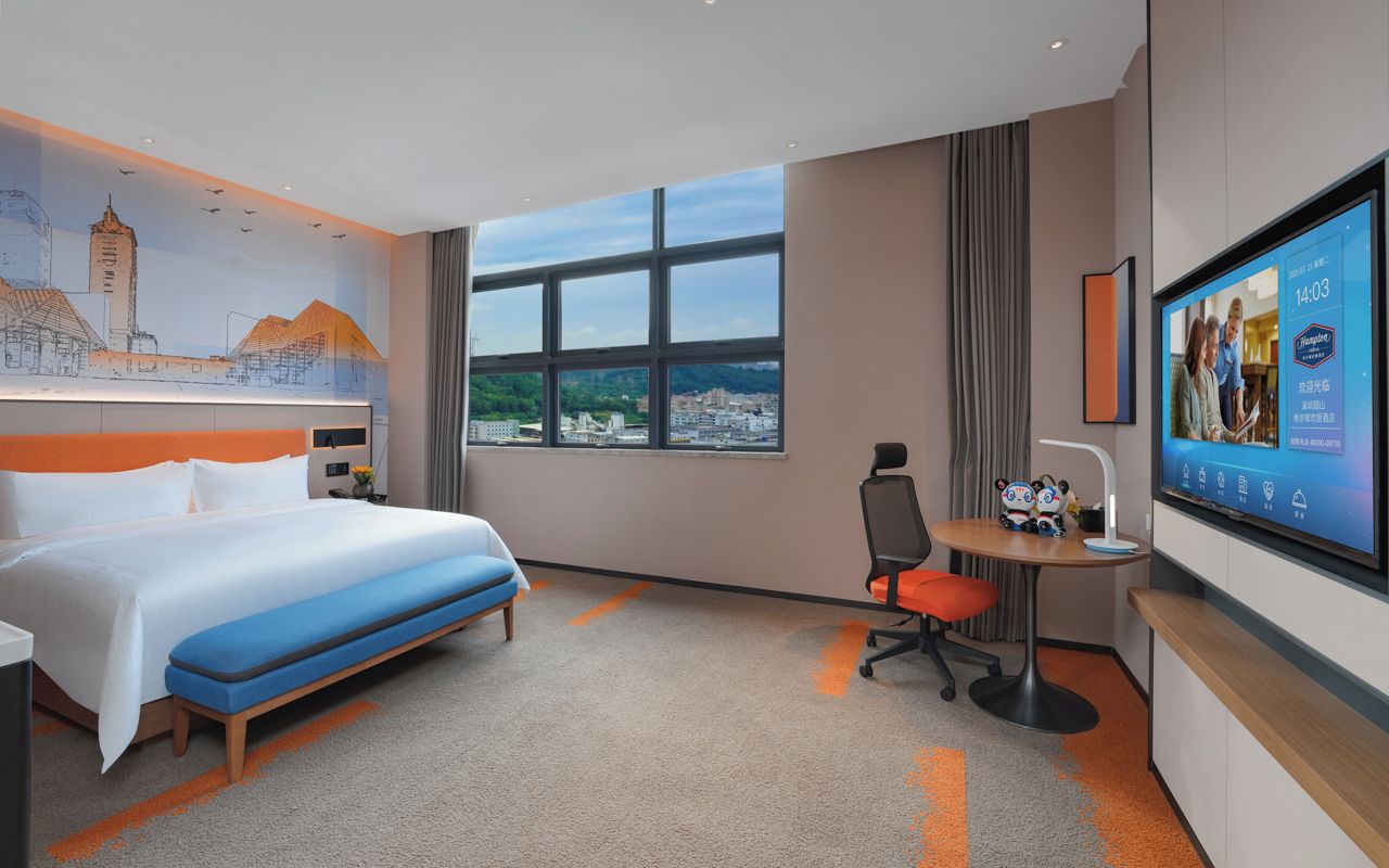 Habitación de hotel con una cama grande, un escritorio, una silla, un televisor y vistas a la ciudad.
