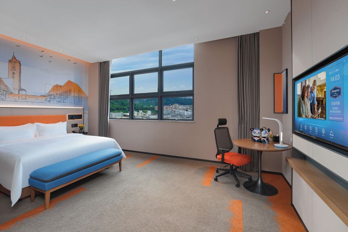 A hotel room with a large bed, a desk, a chair, a television, and a cityscape view.