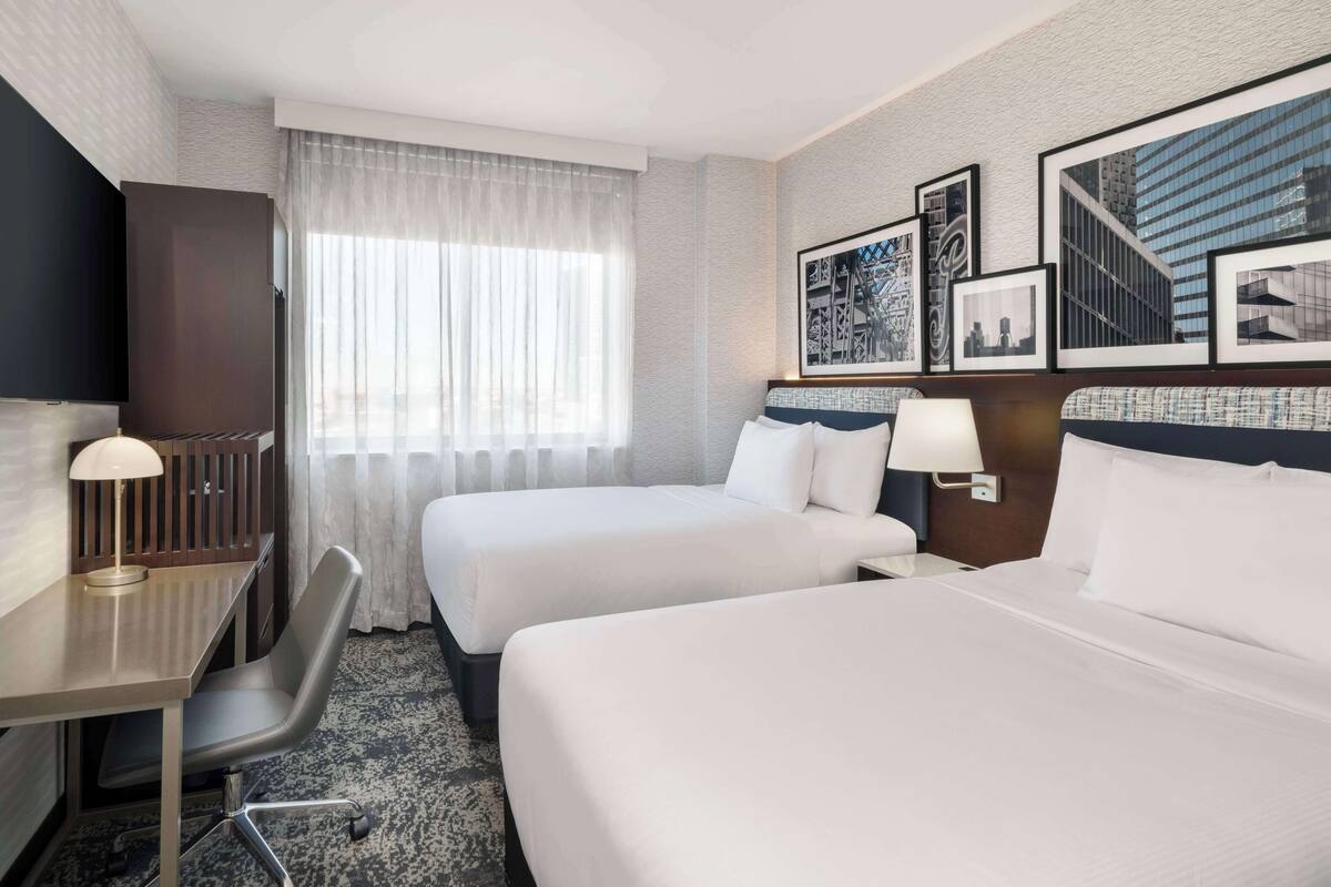Une chambre d’hôtel avec deux lits, un bureau, une chaise et une fenêtre avec des rideaux légers.