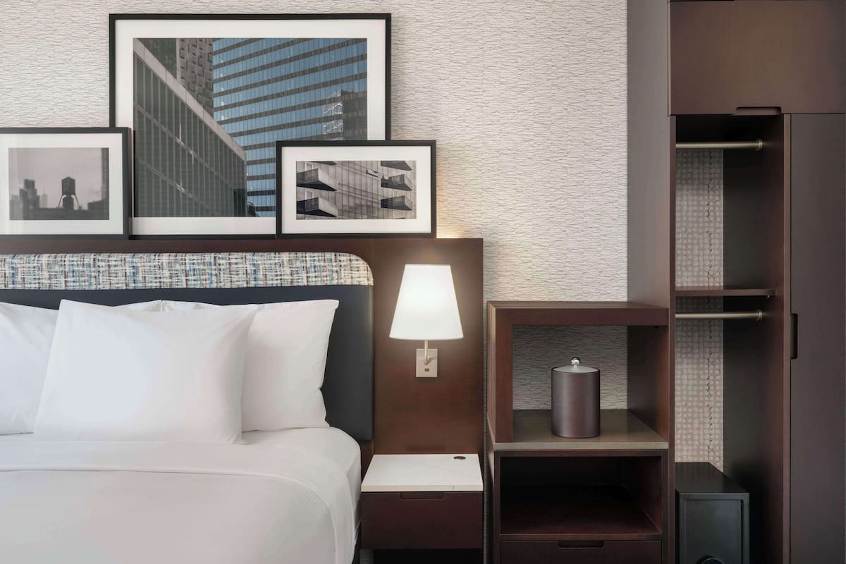 Une chambre d’hôtel comprenant un lit, une table de chevet avec une lampe, une photo encadrée, une petite étagère avec un récipient et une armoire.