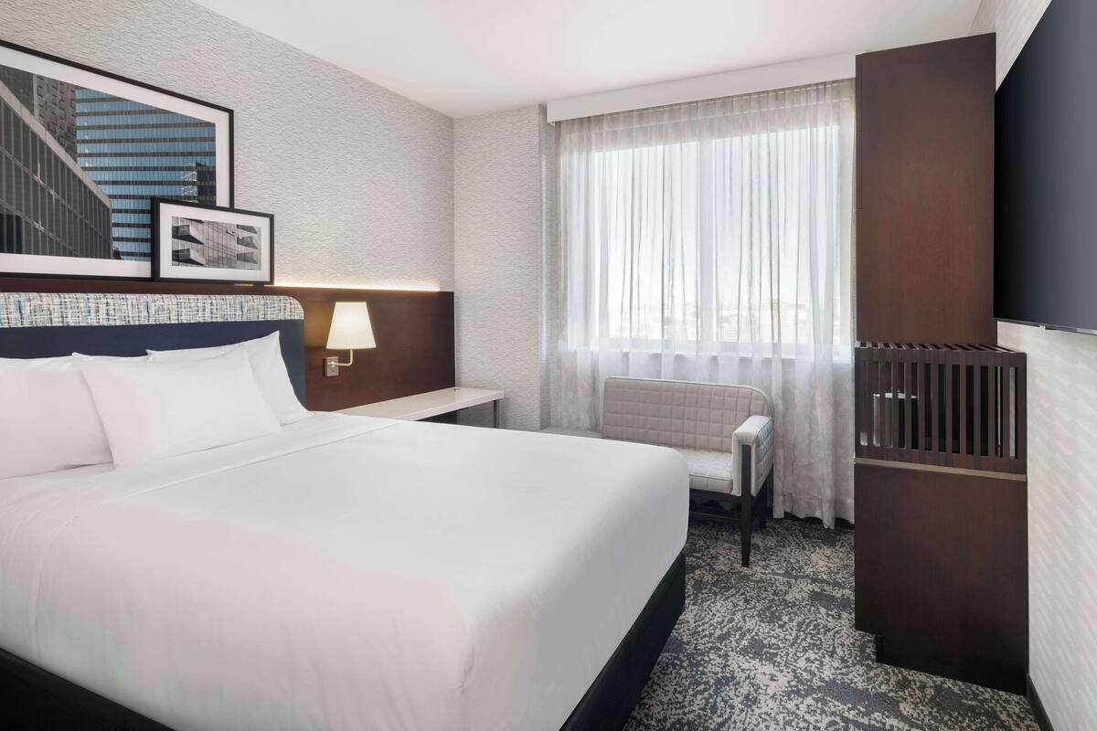 Une chambre d’hôtel avec un grand lit, une table de chevet avec une lampe, une chaise, un téléviseur et une fenêtre avec des rideaux.