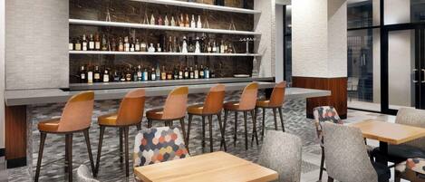 Zona de bar con estanterías llenas de botellas, una barra y asientos con sillas estampadas.