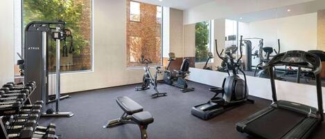A modern gym with various exercise equipment, large windows, and a brick wall.