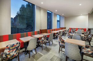 Ein Restaurant mit hohen Tischen, Sitzbänken, gemusterten Stühlen und großen Fenstern mit Blick ins Freie.