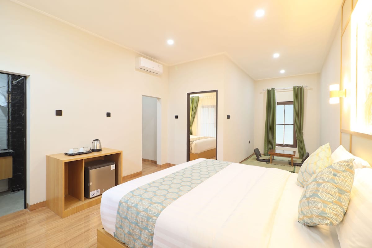 A hotel room with a bed, a desk with a kettle, a mirror, and a window with curtains.