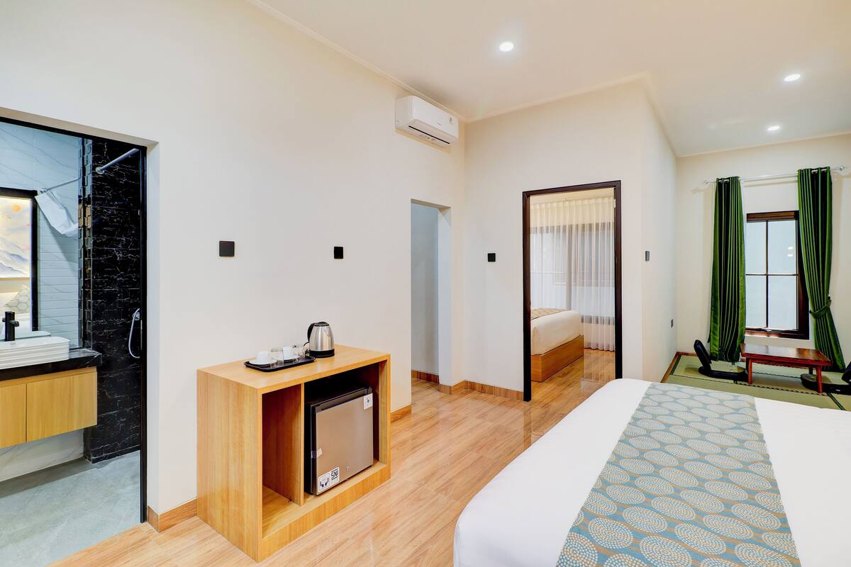 A hotel room with a bed, a small wooden cabinet, a desk, a window with curtains, and a bathroom with a shower and toilet.