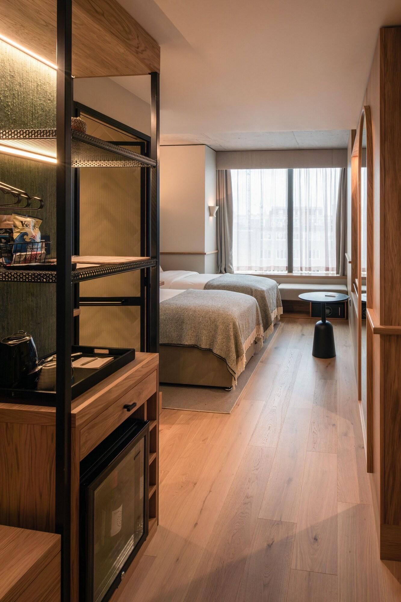 A modern hotel room with a wooden floor, a bed with a headboard, a seating area with a table, and a large window with curtains.