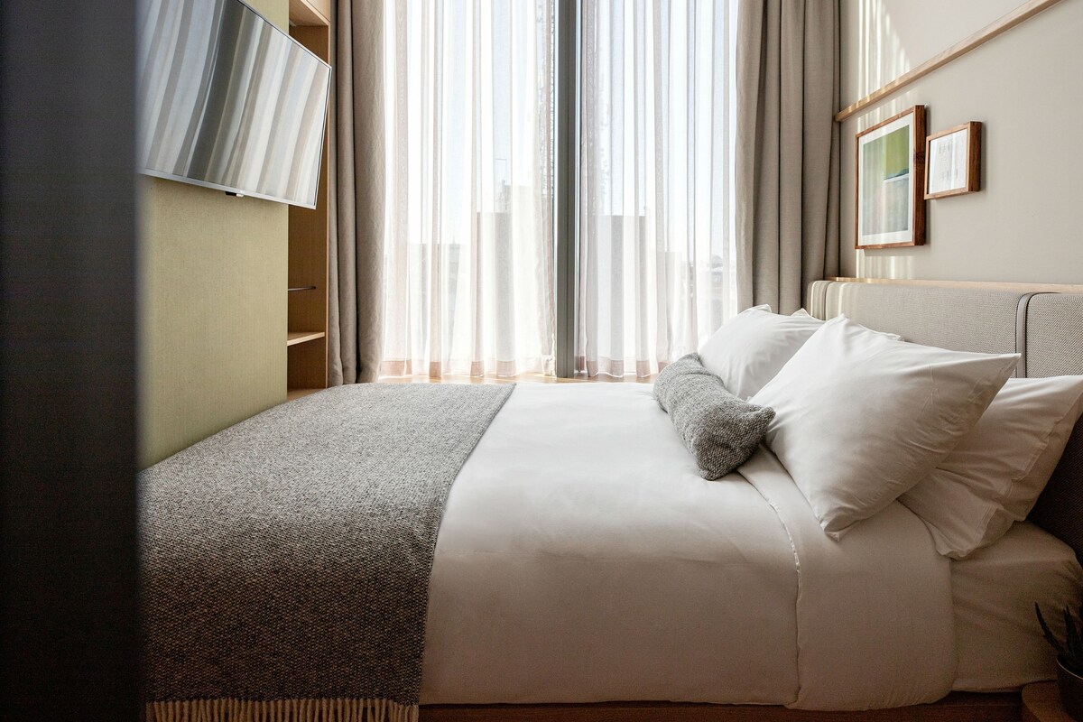 A neatly made bed with white linens and a gray throw blanket, a wooden headboard, and a window with sheer curtains allowing natural light.
