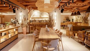 A spacious, modern dining area with a long wooden table and chairs, surrounded by shelves and potted plants.