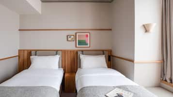 A hotel room with two single beds, a wooden headboard, a small bedside table, a wall-mounted lamp, and a framed picture on the wall.