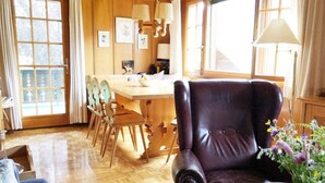 Um quarto aconchegante com paredes de madeira, uma janela grande, uma área de refeições com mesa e cadeiras de madeira, uma poltrona de couro e um vaso com flores sobre uma mesinha lateral.