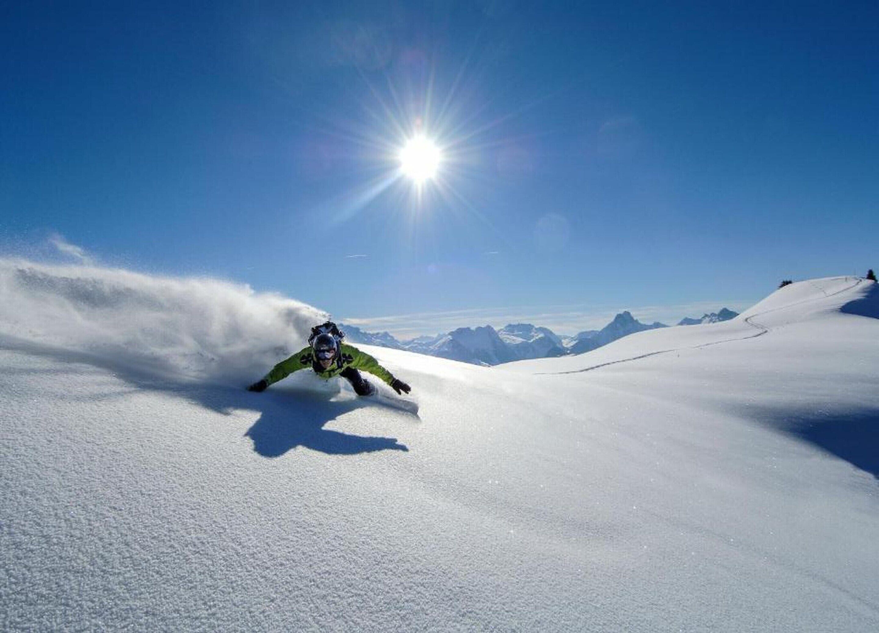 A snowboarder carving a turn on a snow-covered slope with a clear blue sky and distant mountains in the background.