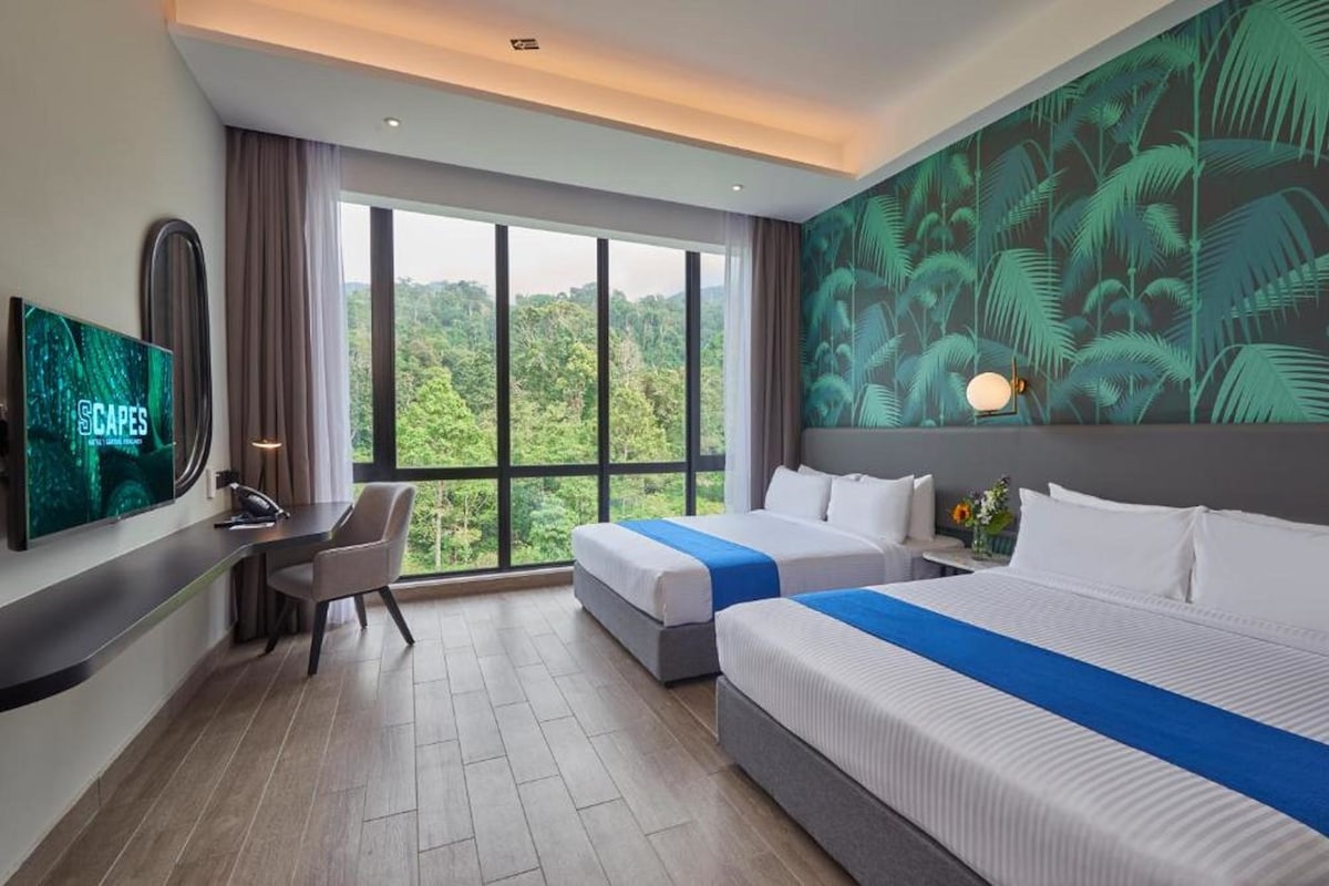 A hotel room with two beds, a desk, a TV, and a large window with a view of the forest.