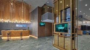 Zona de recepción de hotel con diseño moderno, que incluye un mostrador de recepción, una pantalla digital con el nombre del hotel y una pared decorativa de madera.
