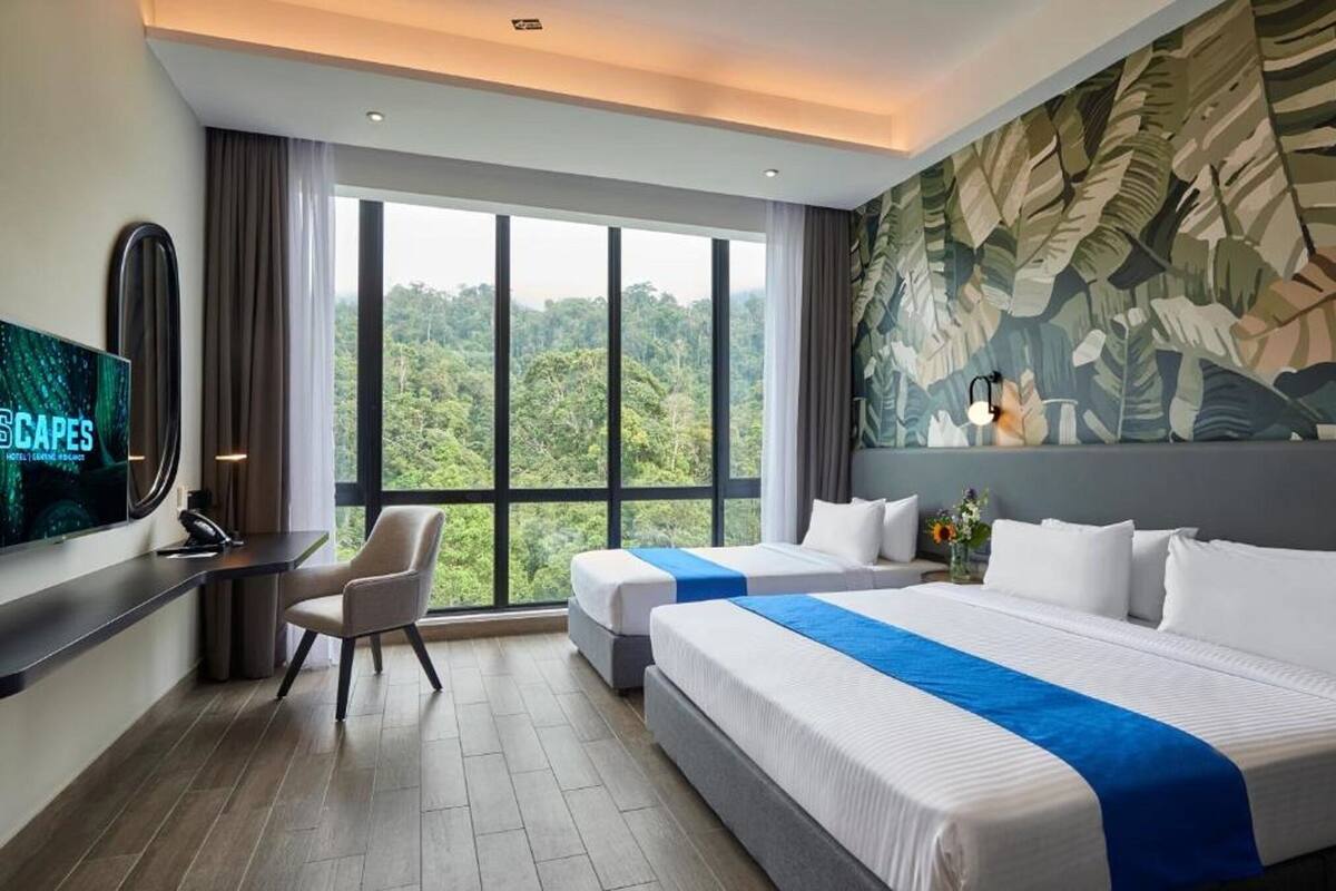 A hotel room with two beds, a desk, a TV, and a large window with a forest view.