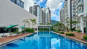 Una piscina rodeada de modernos edificios de apartamentos con balcones, cielo despejado y algo de vegetación.