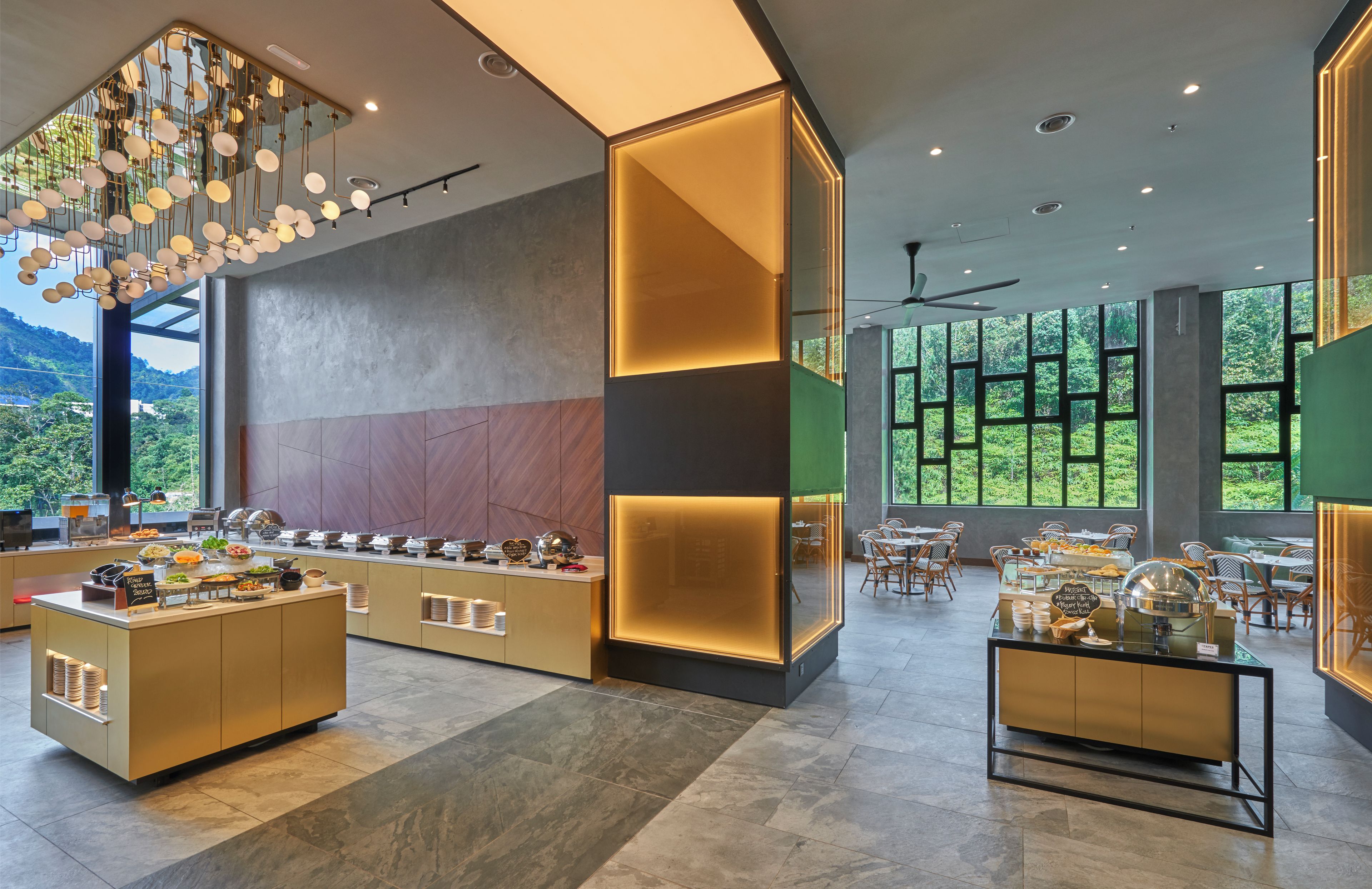 A modern hotel lobby with a buffet area, large windows offering a view of greenery, and contemporary lighting fixtures.