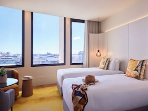 Une chambre d’hôtel avec deux lits, une vue sur la ville, une petite table avec une plante et un luminaire suspendu.