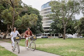 Two people riding bicycles on a sidewalk with trees and a building in the background. - Crystalbrook Kingsley (Newcastle)
