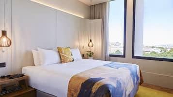 A modern hotel room with a large bed, bedside tables, a nightstand, a window with a city view, and a ceiling light fixture.