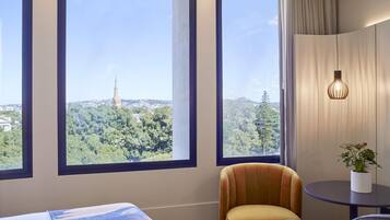 A hotel room with large windows, a bed with a blue and white patterned bedspread, a small round table with a potted plant, and a colorful armchair.