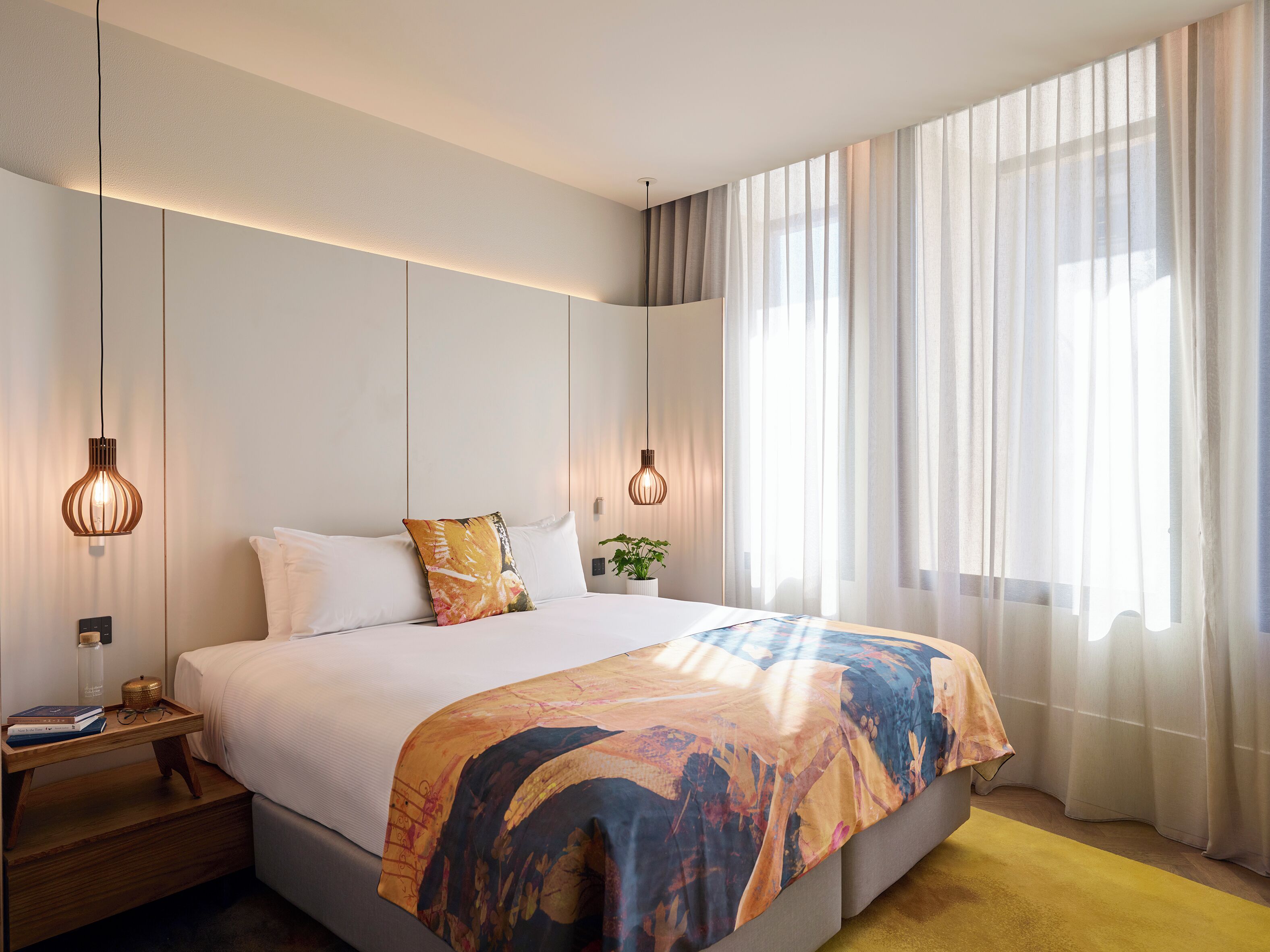A modern hotel room with a large bed, two pendant lights, a bedside table with books, a small plant, and sheer curtains.