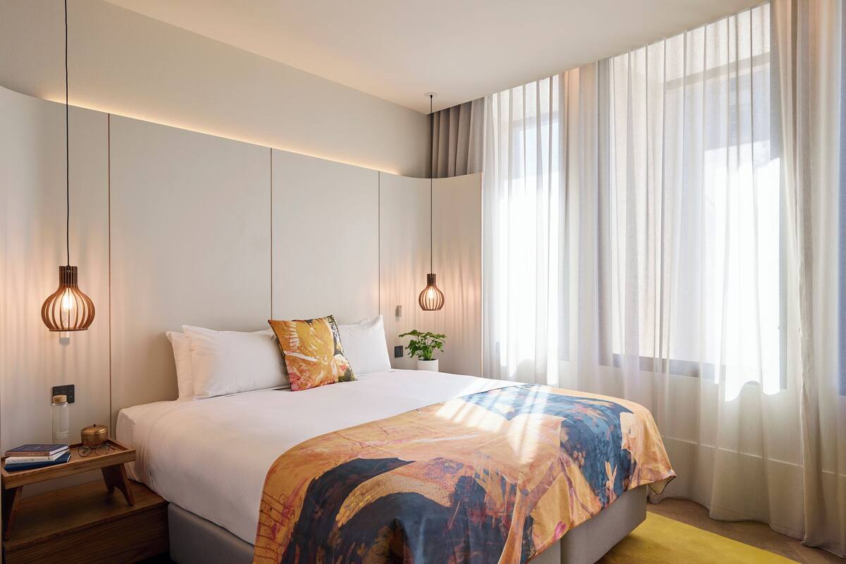 Una camera d'albergo moderna con un letto grande, due lampadari a sospensione, un comodino con una lampada e tende leggere.