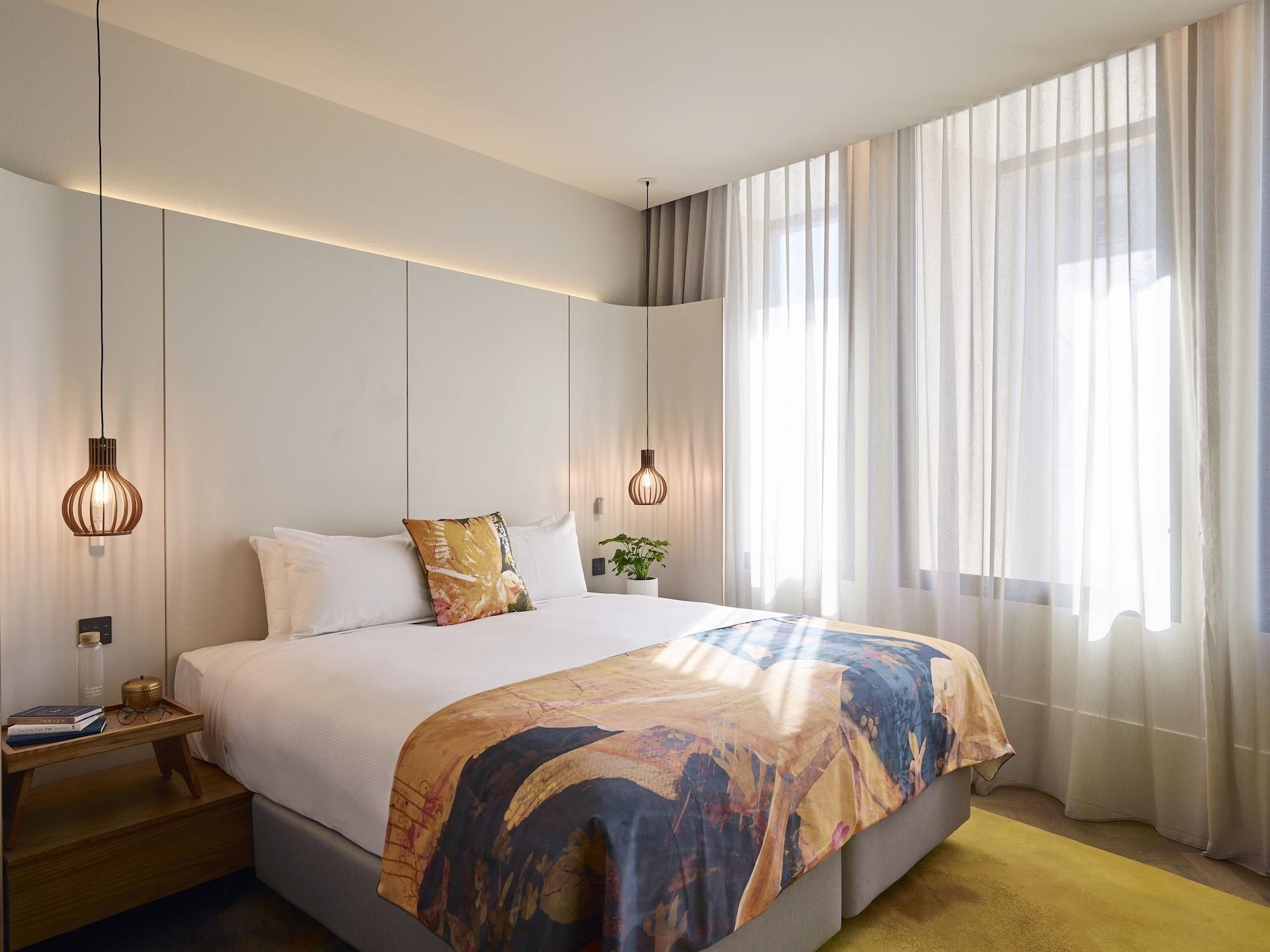 A modern hotel room with a large bed, two bedside lamps, a nightstand, and a window with sheer curtains.