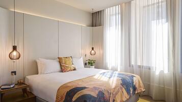 A modern hotel room with a large bed, two bedside lamps, a nightstand, and a window with sheer curtains.