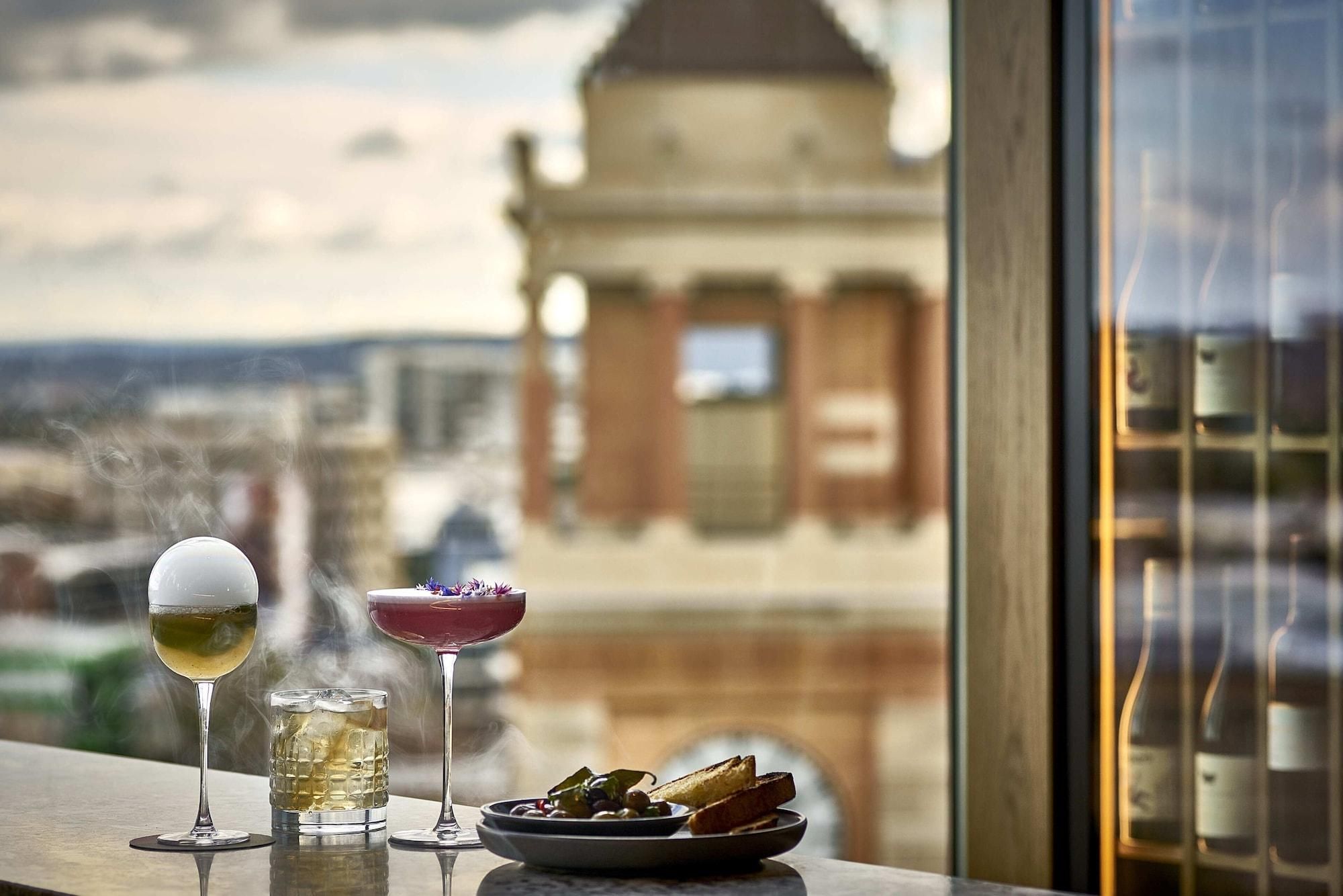 A table with three drinks and a plate of food, overlooking a cityscape with a prominent building.