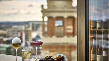 A table with three drinks and a plate of food, overlooking a cityscape with a prominent building.