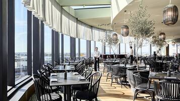 A modern restaurant with high ceilings, large windows, and a view of the city. Tables are arranged for dining, and there are decorative hanging lights.